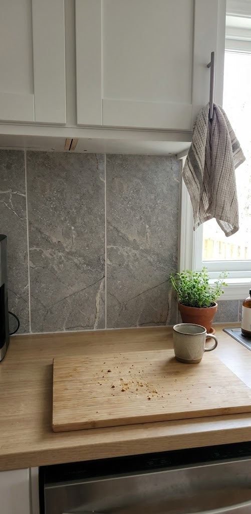 Kitchen counter with a cutting board and mug, stone backsplash, and window with plants.